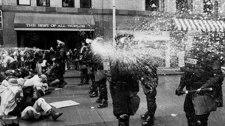 Un oficial de policía de Seattle rocía al fotoperiodista George P. Hickey durante las protestas contra la OMC en Seattle, el 30 de noviembre de 1999. Al fondo, otros oficiales vestidos con equipo antidisturbios también rocían a los manifestantes sentados. Hickey recibió un acuerdo monetario como resultado de haber sido rociado como miembro de la prensa. (George P. Hickey / Sociedad Histórica del Estado de Washington)