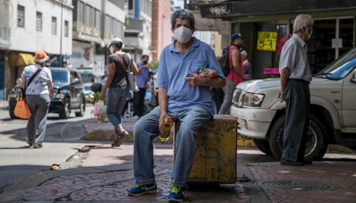 En la imagen, un hombre sostiene una botella de aceite y una bolsa de tomates mientras permanece sentado en una esquina de Caracas. Fotografía: EFE/ Miguel Gutiérrez.